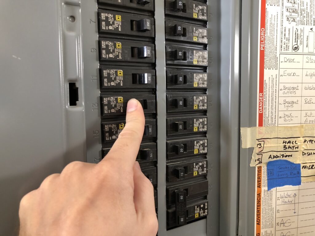 A hand flipping a switch on a circuit breaker panel with labeled breakers and a circuit directory on the right.