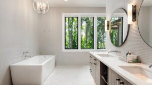 Modern Bathroom With A Rectangular Bathtub, Double Sinks With Round Mirrors, And A Large Window View Of Trees.