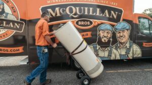 A Man In An Orange Shirt And Jeans Loads A Large Cylindrical Tank Onto A Hand Truck Beside A Mcquillan Bros. Van Featuring Illustrations Of Two Men.