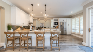 Modern Kitchen With Wooden Bar Stools At An Island Counter, Pendant Lights Above, White Cabinets, And Stainless Steel Appliances. Large Windows With Shutters On The Right Side.