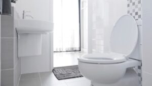 A Bright Bathroom With A White Toilet, Sink, And A Small Rug. The Room Has Tiled Walls And A Window Covered By A Sheer Curtain.