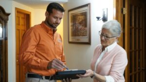 A Man In An Orange Shirt Shows A Tablet To A Woman In A Pink Cardigan Inside A Room With Framed Pictures On The Walls.