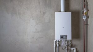 A White Water Heater Is Mounted On A Gray Wall, With Several Pipes Connected Below And A Pressure Gauge On The Right.