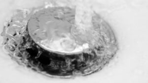 Water Flows Over A Shiny Metal Sink Stopper, Creating Bubbles And Ripples.
