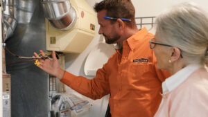 A Technician In An Orange Shirt Explains Electrical Wiring To An Older Woman In A Light-Colored Sweater, Standing Beside Hvac Equipment.