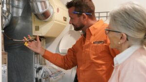 Technician In Orange Shirt Explains Wiring To A Silver-Haired Woman Near Hvac Equipment.