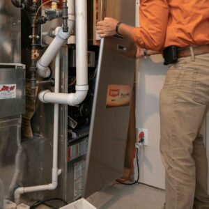 Person In Orange Shirt And Khaki Pants Is Removing The Front Panel Of A Bryant Plus 90 Furnace In A Utility Area With Visible Pipes And Wires.