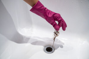 A Hand Wearing A Pink Rubber Glove Pulls A Clump Of Hair From A Bathroom Sink Drain—A Simple Step In Regular Bathroom Drain Maintenance.