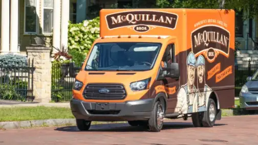 An Orange And Black Mcquillan Bros Service Van, Emblazoned With Company Branding And Vibrant Illustrations, Is Parked On A Residential Street, Ready For Prompt General Heating Repair Services.
