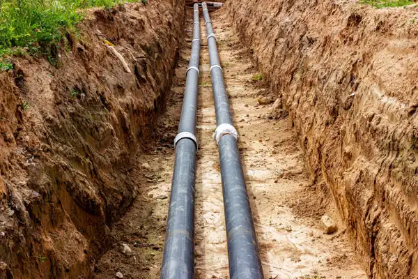 Two Large Black Pipes, Part Of A Sewer Line Installation, Lay Nestled In A Narrow Trench Surrounded By Soil Under A Clear Sky.