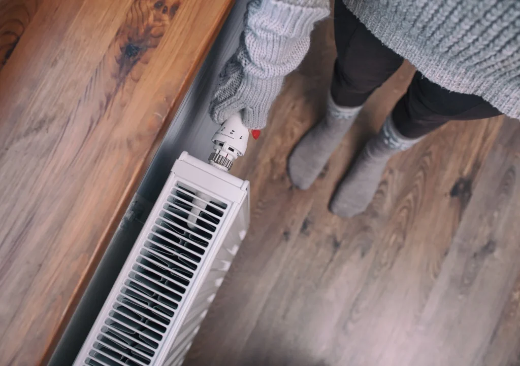 Person in a gray sweater and gloves adjusting the temperature knob on a white radiator, standing on a wooden floor.