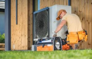 A Technician Wearing An Orange Cap And Tool Belt Repairs Or Maintains An Outdoor Air Conditioning Unit Next To A Wooden Building.