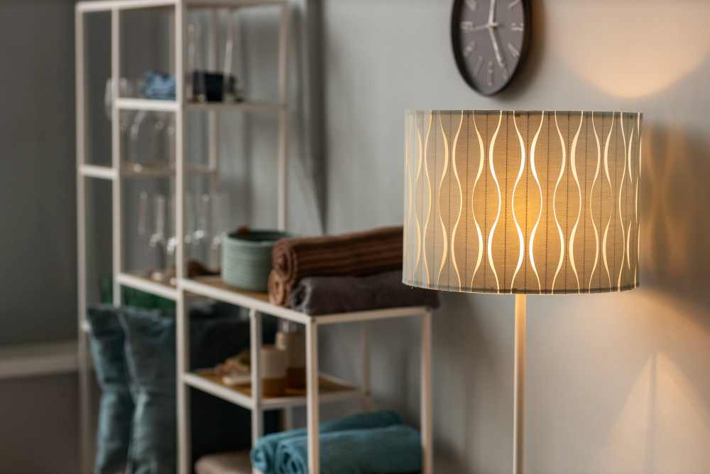 A lit floor lamp with a patterned shade stands near white shelving stocked with towels and glassware; a wall clock hangs in the background.