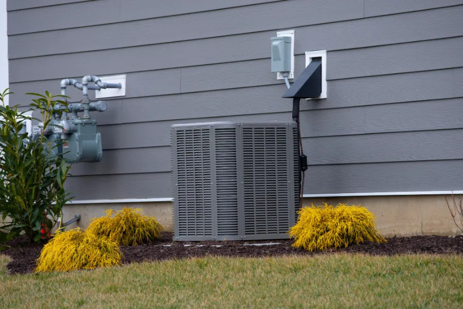An Outdoor Air Conditioning Unit Is Installed Next To A Gray House With Siding, Surrounded By Mulch, Small Shrubs, And Grass.