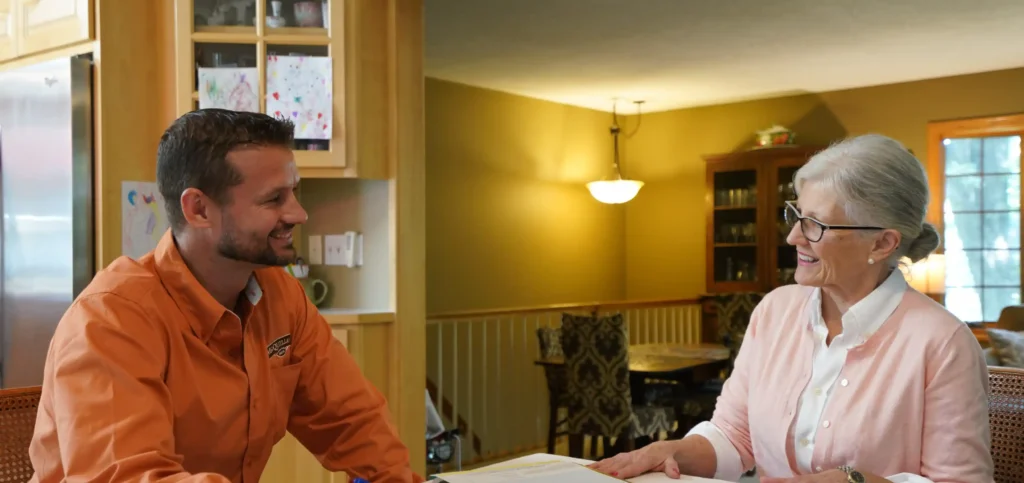 A Man And An Older Woman Sit At A Kitchen Table, Having A Conversation And Looking At Documents Together.