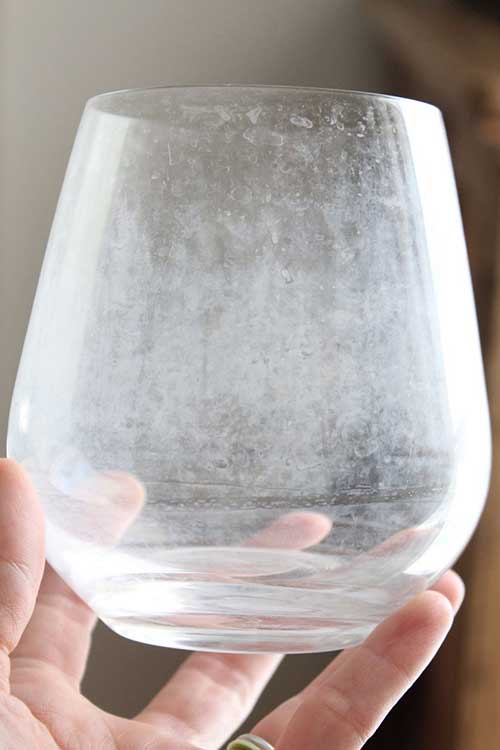 A Hand Holds A Clear Glass With Visible White, Cloudy Hard Water Stains On Its Surface, Highlighting The Effects Of Water Hardness And The Need For A Water Softener System.