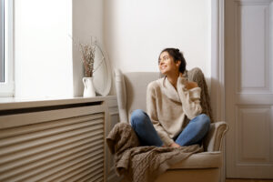 A Woman Sits Cross-Legged On An Armchair, Wearing A Beige Sweater And Jeans, Looking Out A Window. A Vase With Branches Is On The Windowsill Beside Her As She Enjoys The Comfort Of An Efficient Heating And Cooling System.