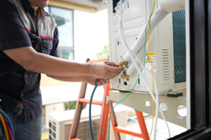 A Technician Adjusts Cables And Pipes Connected To An Outdoor Air Conditioning Unit While Standing On An Orange Ladder.