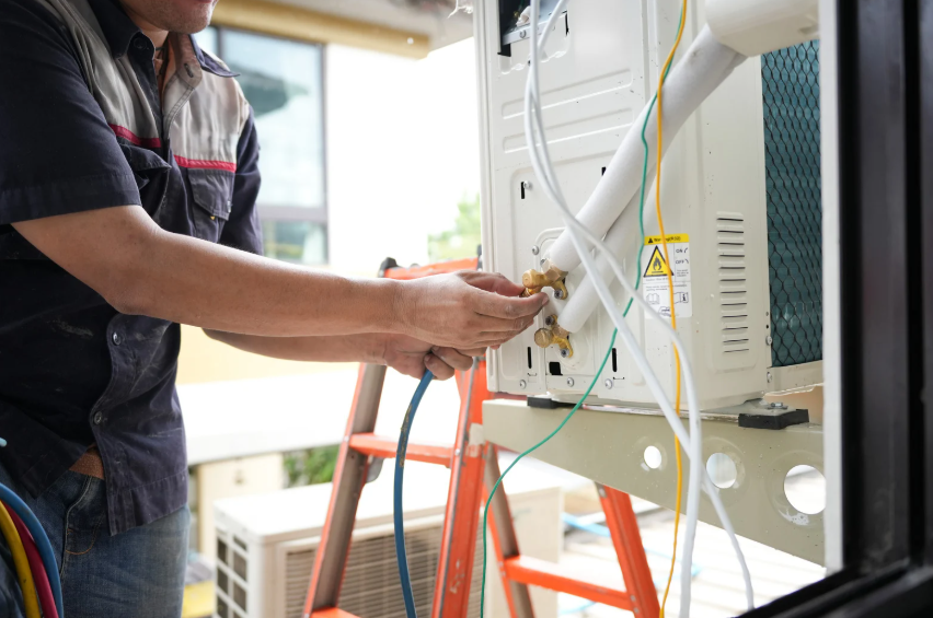 A technician adjusts cables and pipes connected to an outdoor air conditioning unit while standing on an orange ladder.