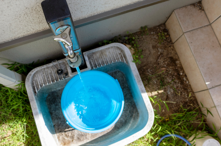 A Blue Bucket Is Being Filled With Water From An Outdoor Faucet, Placed In A Gray Basin Near Tiled Steps And Green Grass.