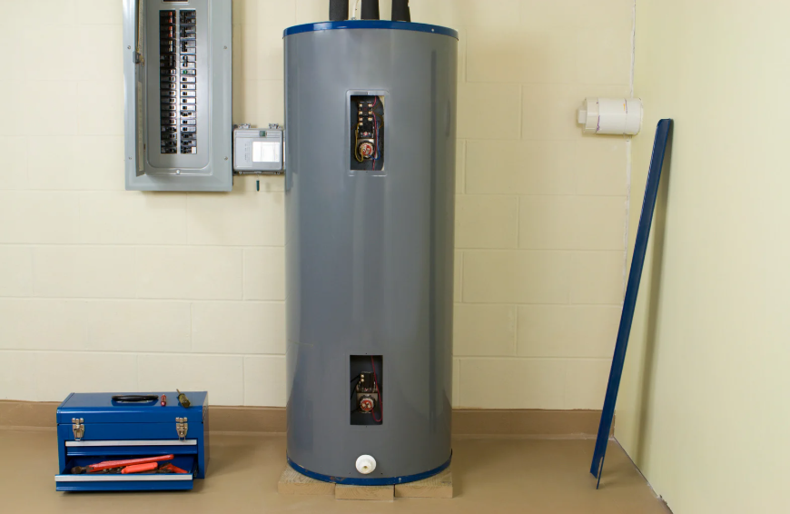 A gray electric water heater stands next to a circuit breaker panel on a beige basement floor, with a blue toolbox and tools nearby.