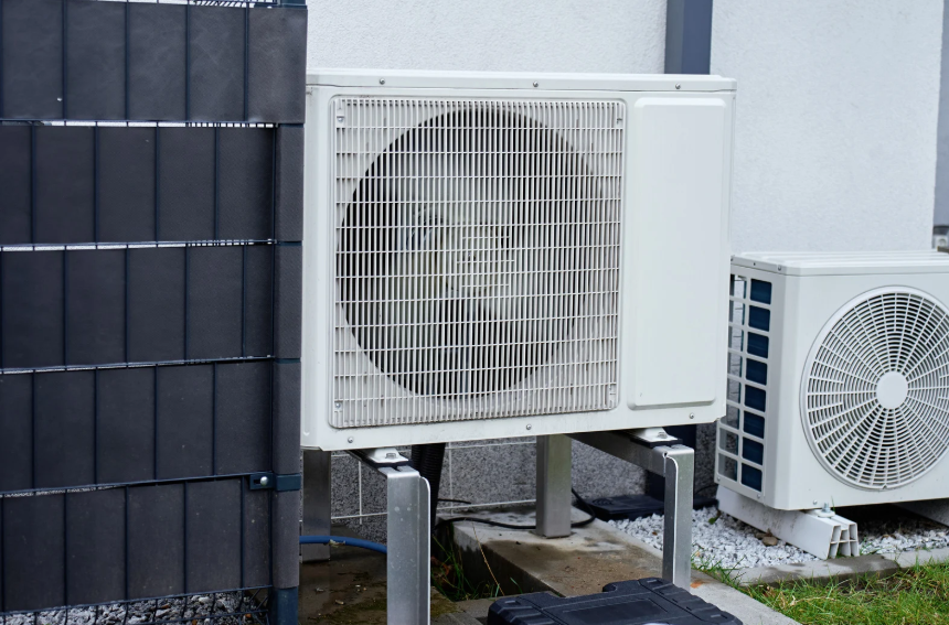 Two Outdoor Hvac Units Are Installed On Metal Stands Next To A Building, With A Black Fence Panel And Some Grass Visible Nearby.