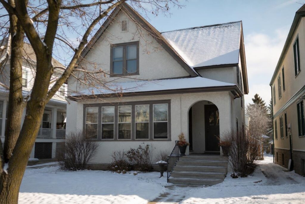 Snow-Covered Single-Family House With An Arched Front Porch, Leafless Tree In Front Yard, And Neighboring Homes Visible On Both Sides; Residents May Experience Low Water Pressure In The House During Winter Months.