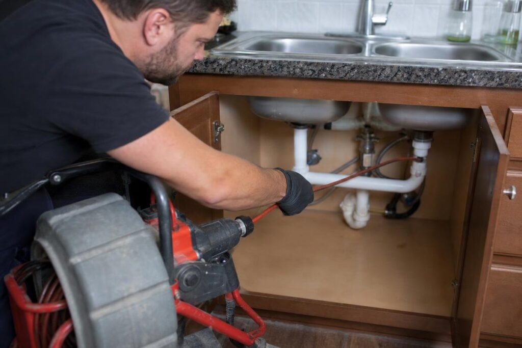 A Person Uses A Plumbing Snake To Unclog Pipes Under A Kitchen Sink With Double Basins And Brown Cabinets, Addressing Issues That Could Be Causing Low Water Pressure In The House.