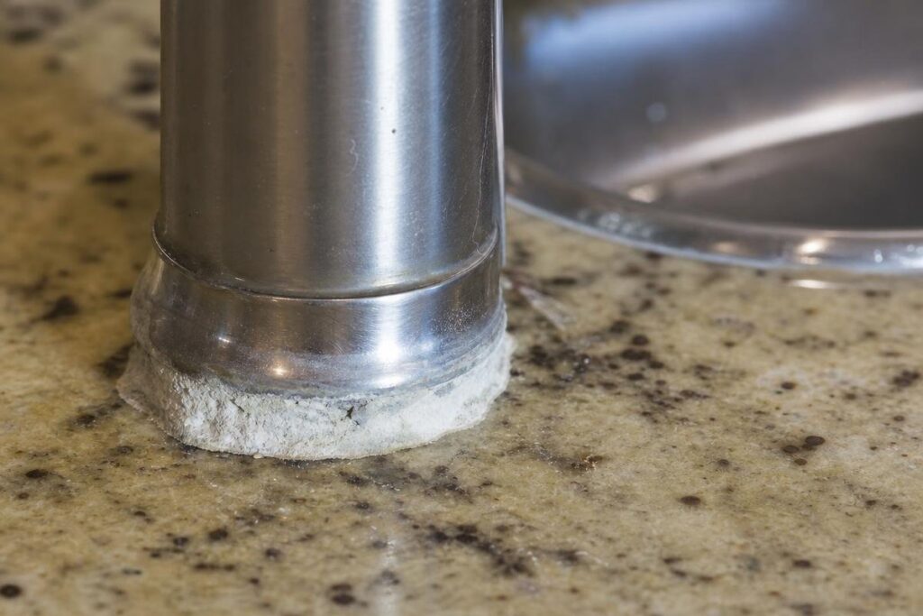 Close-up of a kitchen faucet base with white, chalky mineral buildup on a granite countertop near a stainless steel sink, which can sometimes be a sign of low water pressure in house.