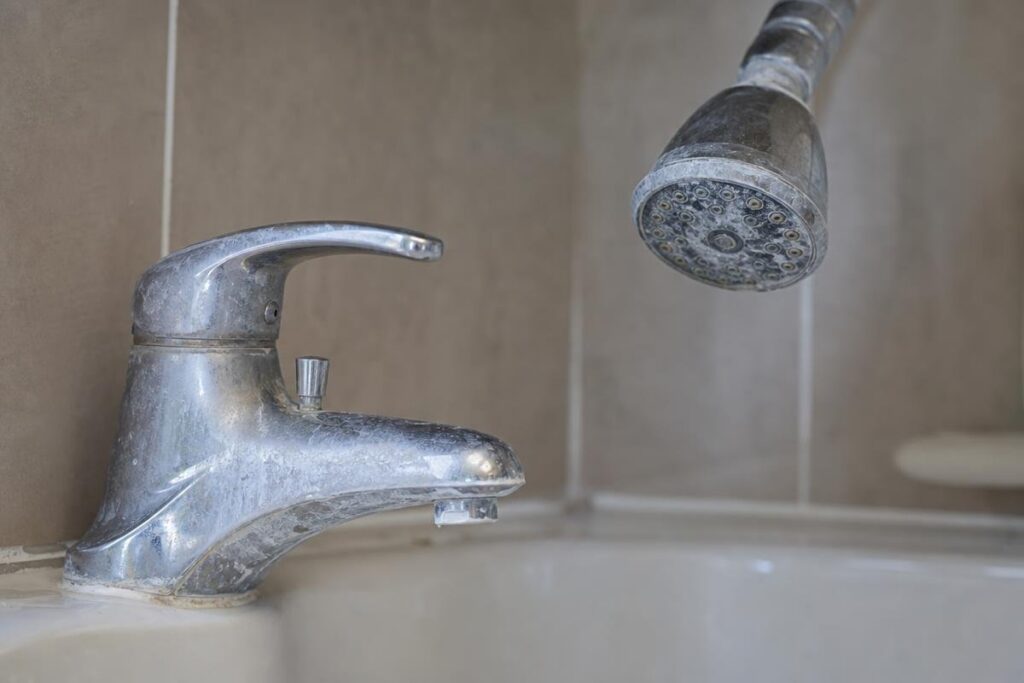 Close-Up Of A Bathtub Faucet And Showerhead With Visible Limescale And Hard Water Stains, Set Against Beige Tiled Walls—Common Signs That Can Contribute To Low Water Pressure In House.