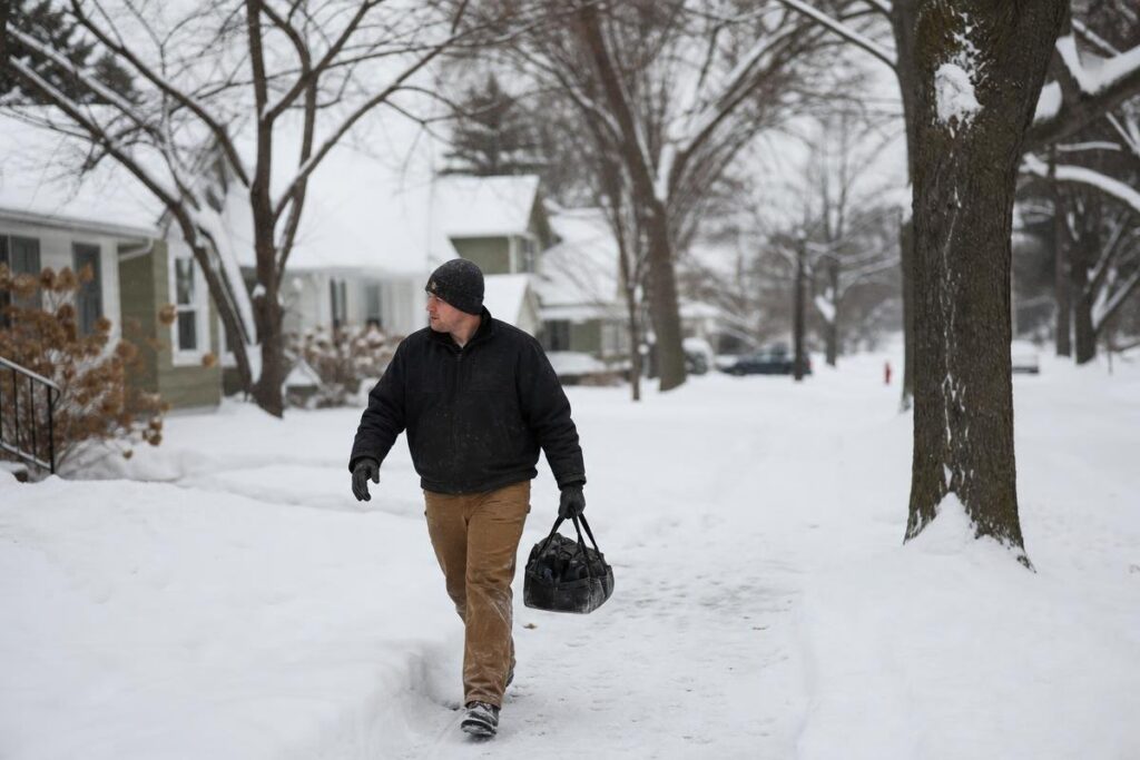 A Man Walks On A Snowy Sidewalk In A Residential Neighborhood, Carrying A Black Duffel Bag And Wearing Winter Clothing. As He Passes By Snow-Covered Homes And Frosted Trees, He Thinks About The Low Water Pressure In His House.