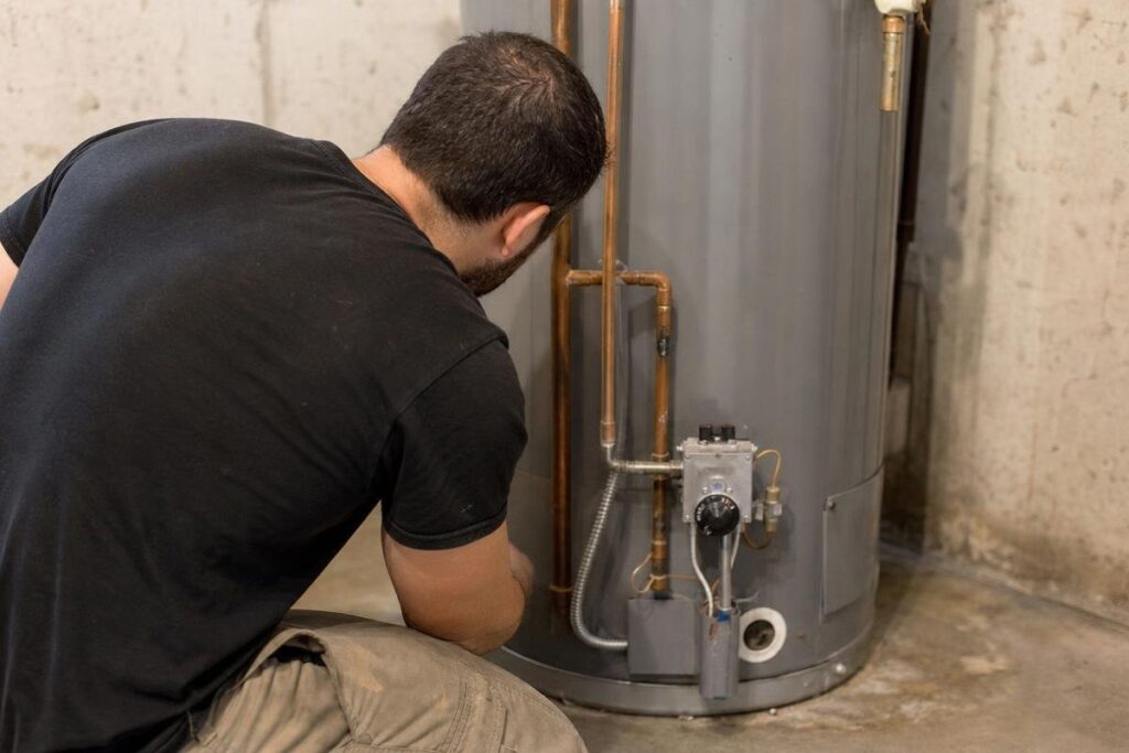 A person kneels and inspects the pipes and controls at the bottom of a gray water heater in a basement with concrete walls, possibly investigating low water pressure in the house.