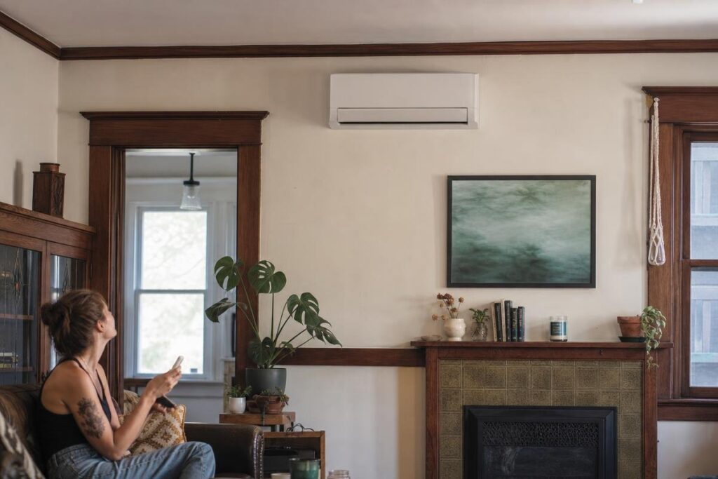 A person sits on a couch in a living room with a wall-mounted air conditioner above a fireplace, a plant, and a painting on the wall, perhaps contemplating recent issues like low water pressure in the house.