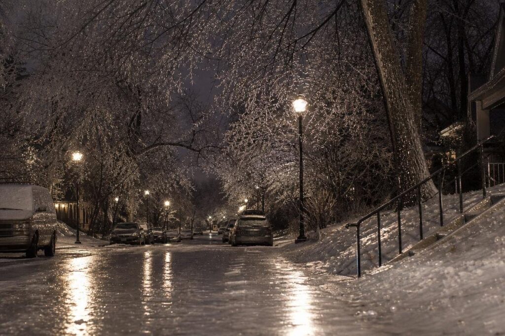 A Residential Street At Night Is Covered In Ice, With Parked Cars And Trees Coated In A Shimmering Layer Of Ice, Illuminated By Streetlights. The Quiet Stillness Is Broken Only By The Occasional Concern From Nearby Homeowners About Low Water Pressure In The House, A Common Issue After Winter Storms Like This One.