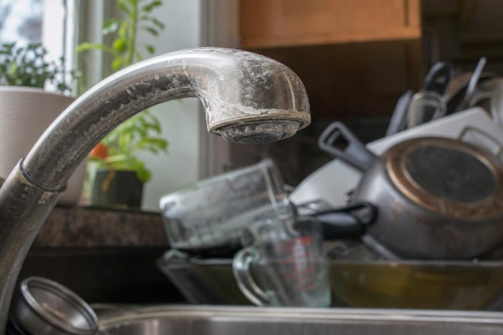 A Kitchen Faucet With Mineral Buildup Is In The Foreground, A Possible Sign Of Low Water Pressure In The House, While A Pile Of Dirty Dishes, Pots, And Glassware Is Stacked In The Sink Behind It.