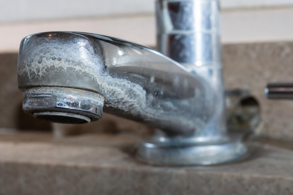 Close-Up Of A Metal Faucet With Visible White Limescale And Mineral Deposits On Its Surface, Installed On A Brown Countertop—Common Signs That May Contribute To Low Water Pressure In House Settings.